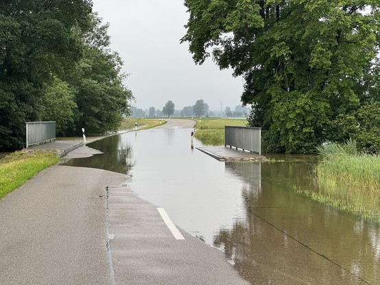 A flooded rural road near Donauwoerth surrounded by trees and grass, with water covering the pavement and part of the sidewalk.