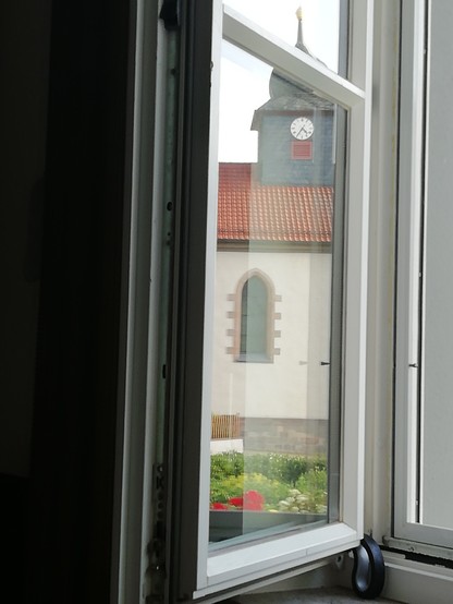 Ein geöffneter Fensterflügel spiegelt den Turm der gegenüber liegenden Kirche. Die Turmuhr zeigt 7.25 Uhr an (gespiegelt 16.35 Uhr). Das Dach ist mit roten Dachpfannen gedeckt. Im Vordergrund Ausschnitt eines bunten Blumenbeetes.