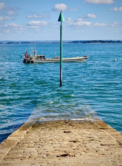 A stone boat ramp leading into the water, with a green navigation marker in the foreground and a boat in the distance. The sky is clear with a few clouds. / @infoleck