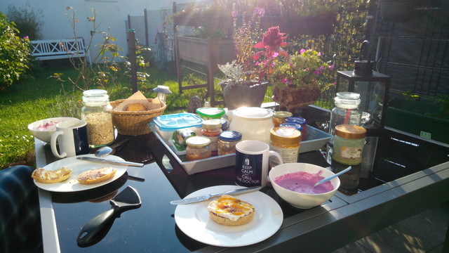 Blick auf einen gedeckten Terassentisch mit Müslischälchen und Brötchen, und Kaffee, versteht sich. Im Hintergrund ein Reihenhausgarten mit Wiese und Büschen. Sonnenschein: Niedrig stehende Herbstsonne von rechts. 