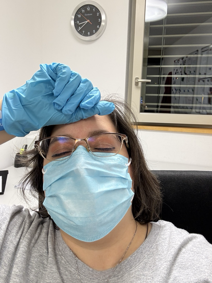 kurze Verschnaufpause im Stationszimmer; Selfie, mit rechter Hand in blauem Silikonhandschuh auf fiebriger Stirn, Augen geschlossen, Medizinmaske tragend. Im Hintergrund ein Fenster und eine schwarze Wanduhr.