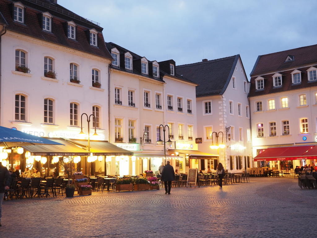 Blick auf einen Teil des St. Johanner Marktes in Saarbrücken. Mehrere alte helle Häuser bilden  eine Ecke. Vor den Häusern ist beleuchtete Außengastronomie mit teilweise bunten Markisen und Marktschirmen.